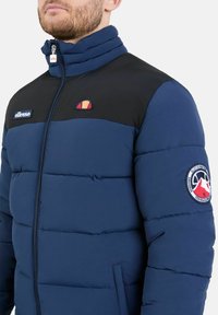 Giacca imbottita blu navy con una sezione superiore nera. Presenta un alto collo, una cerniera e loghi sul petto e sulla manica. Texture morbida e liscia.