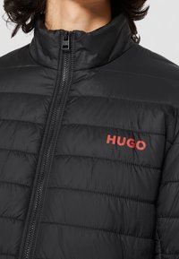 Czarna ocieplana kurtka z wysokim kołnierzem, zamek błyskawiczny z przodu i poziomym przeszyciem; posiada czerwone logo "HUGO" na lewej piersi.