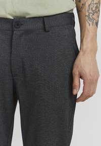 Pantalones grises hechos de una tela texturada, con cierre de botón, bolsillos laterales y un corte a medida. Mano visible cerca de la cinturilla.