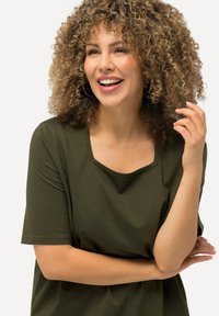 Olivově zelená košile s krátkým rukávem a čtvercovým výstřihem, vyrobená z měkké látky. Modelka má kudrnaté vlasy a lehce gestikuluje rukou.