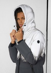 Gewatteerd jack met een wit bovenstuk dat overgaat in een donkergrijs onderstuk, voorzien van een capuchon, ritsen en een zwart cirkelvormig accent op de mouw.