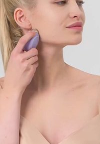 Massager facial portable violet tenu contre une joue, surface lisse, forme arrondie, avec un accent argenté près du bas. Éclairage doux.