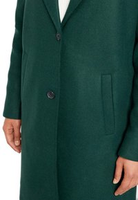 Grüner Wollmantel mit Reverskragen, schwarzer Knopfverschluss und Seitentaschen. Strukturierter Stoff mit glatter Oberfläche.