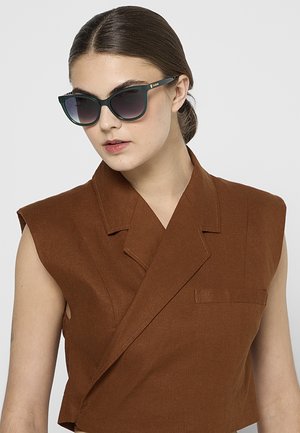 Kvinde med mørkt hår bundet tilbage iført mørkegrønne katteøje-solbriller og en ærmeløs brun blazer med brede revers.