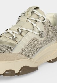 Basket beige épaisse avec tissu moucheté, lacets blancs épais à motifs, œillets métalliques et un pendentif logo rond en argent sur le côté.