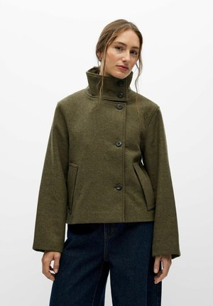 Une femme porte un manteau vert olive boutonné à col montant et un jean foncé, sur un fond blanc uni.