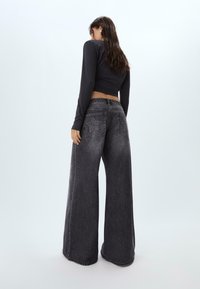 Frau trägt weit geschnittene schwarze Jeans mit Stern-Patches auf den hinteren Taschen und ein figurbetontes schwarzes Langarm-Crop-Top, steht vor einem schlichten Hintergrund.