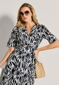 Schwarz-weiß gemustertes Kleid mit V-Ausschnitt und kurzen Ärmeln, tailliert in der Taille, kombiniert mit übergroßen Sonnenbrillen und einer kleinen geflochtenen Tasche.