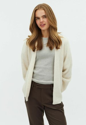 Cardigan a coste color crema con apertura frontale, indossato sopra un maglione grigio chiaro. Il modello indossa pantaloni tagliati marroni scuri. Dettagli del tessuto testurizzato visibili.