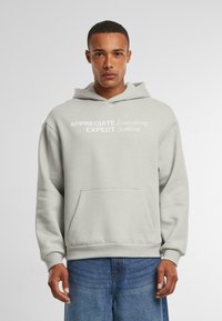 Lichtgrijze hoodie van een katoenmix, met een voorvak en witte bedrukte tekst: "APPRECIEER Alles VERWACHT Niets."