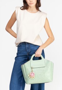Myntgrøn håndtaske med blomstret prægdesign, top-hank og en pink blomstercharms hængende ved. Model iført cremefarvet ærmeløs top og blå jeans.