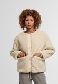 Kobieta z kręconymi włosami ubrana w beżową, z guziczkami, teksturowaną kurtkę polarową z kieszeniami, stojąca na jednolitym jasnym tle.