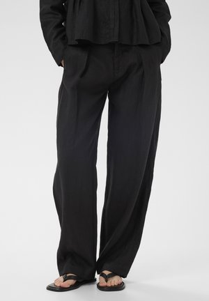 Személy laza fekete lenvászon nadrágot visel, kezét a zsebében tartja, fekete lapos pántos szandált hord egy egyszerű, világos háttér előtt.