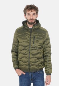 Giacca imbottita verde oliva con zip frontale, cappuccio e motivo a onde testurizzate. Presenta una tasca con zip e polsini elasticizzati. Indossata con jeans blu.