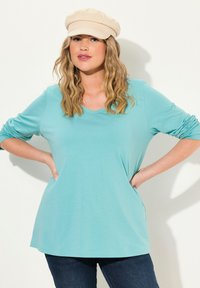 Helles blaues V-Ausschnitt Langarmshirt mit umgeschlagenen Ärmeln, aus weichem Stoff, kombiniert mit einem beigen Hut, getragen mit dunklen Jeans.