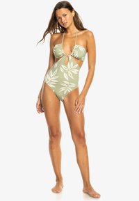 Olijfkleurig eendelig badpak met wit loofpatroon, voorzien van een V-hals, uitsnijding bij de taille en dunne verstelbare bandjes.