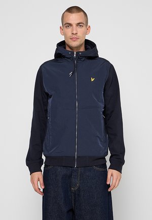 Uomo che indossa una felpa con cappuccio blu navy con zip e logo giallo di uccello e jeans scuri in denim, in piedi su sfondo grigio uniforme.