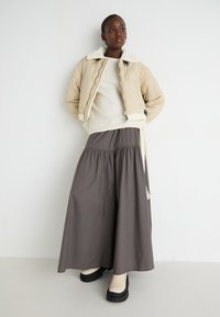 Beige prošívaná bomber bunda s bílým koženým límcem, vrstvená přes světly svetr, spárovaná se šedou sukní po kotníky a masivními černými botami.