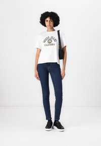Bílý bavlněný tričko s potiskem "Guess Jeans California" v černé a zlaté barvě. Kombinováno s modrými skinny džíny a černými teniskami. Nastavitelná taška.
