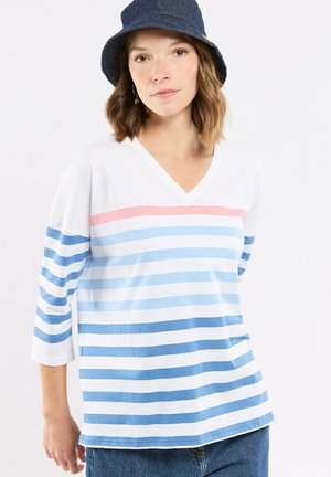 Femme portant un t-shirt blanc à col en V avec des rayures horizontales roses et de différentes nuances de bleu, manches trois-quarts, chapeau seau en jean bleu et jean.