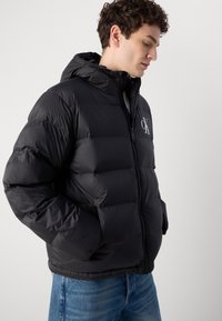 Schwarze Puffjacke mit Kapuze, ausgestattet mit einem Reißverschluss und elastischen Bündchen, leicht gepolstert mit einem sichtbaren Logo auf der Brust.