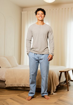 Glunderende man zonder schoenen in een grijs langemmouw shirt en blauwe pyjamabroek staat in een slaapkamer met beige beddengoed en een houten bank.