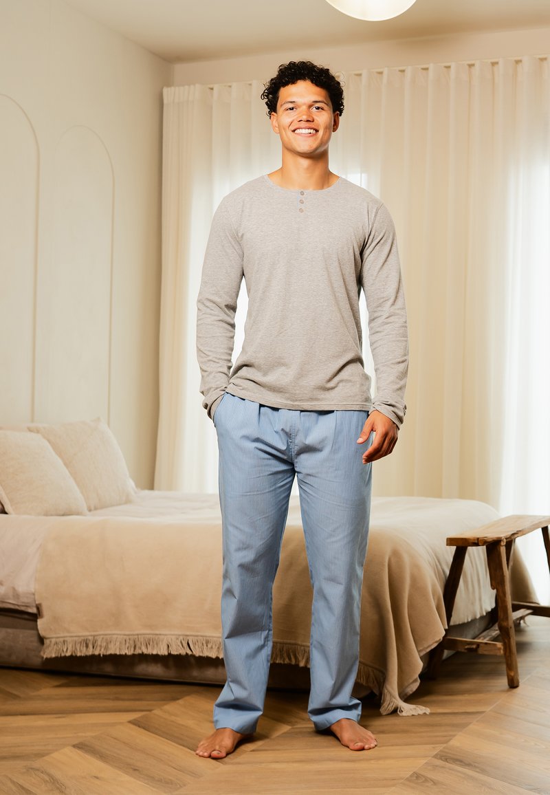 Homme souriant, pieds nus, portant une chemise grise à manches longues et un pantalon de pyjama bleu, se tient dans une chambre avec une literie beige et un banc en bois.