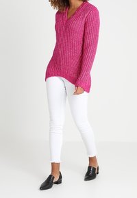 Maglione a maglia fucsia con scollatura a V e due bottoni, abbinato a jeans skinny bianchi e mule nere basse. Presenta polsini e orlo a coste.