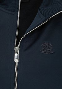 Veste marine zippée avec texture côtelée, fermeture éclair argentée et logo embossé sur la poitrine. Le matériau semble lisse et décontracté.