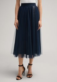 Jupe en tulle bleu marine avec une couche transparente, présentant une large ceinture texturée et un design longueur cheville. Portée avec des talons noirs à brides.