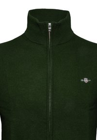 Suéter de cremallera en verde oscuro con cuello alto, textura acanalada y cremallera plateada. Presenta un pequeño logotipo en la parte izquierda del pecho.