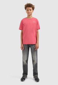 Teenager mit lockigem Haar, der ein korallenfarbiges T-Shirt, abgewetzte schwarze Jeans und schwarze Sneakers trägt und vor einem einfarbigen hellgrauen Hintergrund steht.