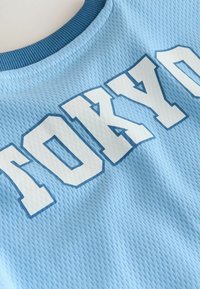 Lyseblåt mesh-stof med fedt hvidt "TOKYO" tekst og mørkeblå konturer. Rund, struktureret krave tilføjer detalje.