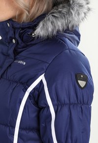 Femme portant une veste d'hiver matelassée bleu marine avec un bord en fausse fourrure grise sur la capuche et des rayures verticales blanches sur les manches.