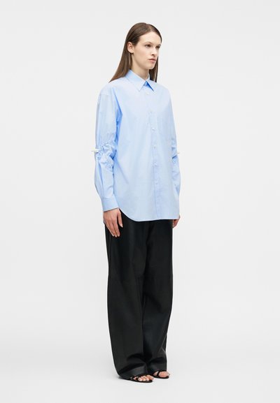 Γυναίκα που στέκεται φορώντας άνετο ανοιχτό γαλάζιο πουκάμισο με κουμπιά και μαζεμένα μανίκια, μαύρο παντελόνι με φαρδιά πόδια και μαύρα πέδιλα ανοιχτά στα δάχτυλα, μπροστά σε απλό φόντο.
