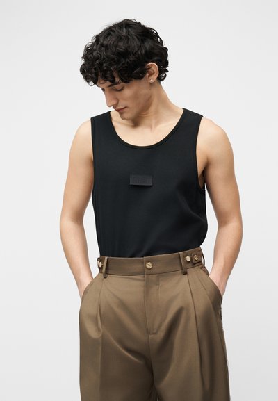 Νεαρός άνδρας με σγουρά σκούρα μαλλιά, φορώντας μαύρο αμάνικο μπλουζάκι και καφέ παντελόνια ψηλής μέσης, με τα χέρια στις τσέπες, κοιτάζοντας προς τα κάτω.