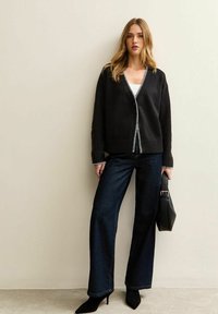 Czarny kardigan z białymi wykończeniami, w połączeniu z ciemnoniebieskimi dżinsami o wysokim stanie i czarnymi szpilkami. Modelka trzyma czarną torebkę.