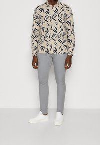 Camisa beige de manga larga con estampado de hojas negras, combinada con pantalones ajustados gris claro y zapatillas blancas. Conjunto casual en general.