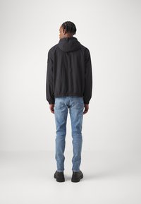 Giubbotto leggero nero con cappuccio, polsini ed orlo elasticizzati, abbinato a jeans in denim azzurro chiaro e scarpe nere. Vista da dietro.