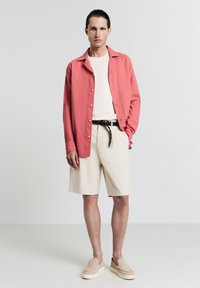 Chemise boutonnee corail, t-shirt blanc, shorts crème, ceinture tissée noire, et chaussures à enfiler beiges. Le mannequin se tient devant un fond neutre.