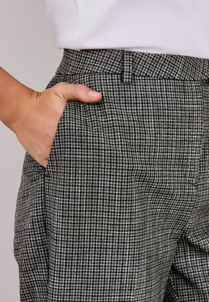 Mână odihnindu-se în buzunarul din față al pantalonilor în carouri alb-negru purtați cu o cămașă albă băgată în pantaloni.