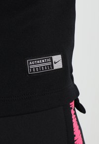 Schwarze Sportkleidung mit einem pinken abstrakten Muster an der Seite, versehen mit einem Etikett mit der Aufschrift "AUTHENTIC FOOTBALL" und Nike-Branding.