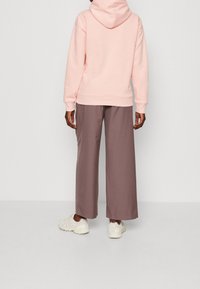 Sudadera con capucha de color rosa claro y corte relajado, combinada con pantalones morados de pierna ancha y zapatillas blanco crema. La textura parece suave.