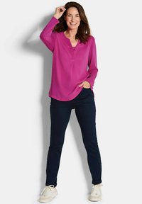 Blusa magenta a maniche lunghe con colletto a V, abbinata a pantaloni blu scuro e sneakers bianche. La blusa ha una texture liscia e una vestibilità comoda.