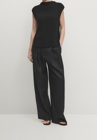 Zwart geribd gebreid mouwloos top met hoge hals, gecombineerd met losse zwarte broek met plooien en een gladde textuur. Zwarte sandalen.