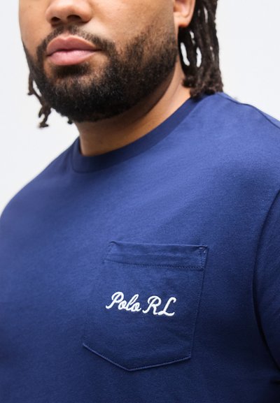 Κοντινό ενός άνδρα που φοράει μπλε μπλούζα με μικρό τσεπάκι στο στήθος, κεντημένο με τη φράση "Polo RL" σε λευκό νήμα.