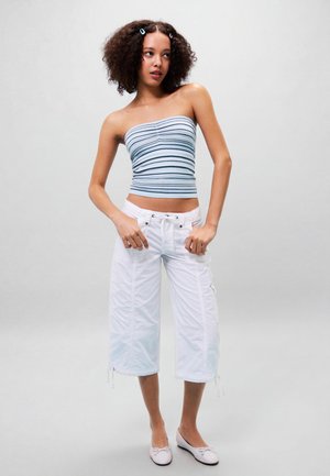 Fiatal nő hullámos hajjal, aki csíkos tube topot, fehér rövidített nadrágot és világos rózsaszín lapos cipőt visel, egy egyszínű háttér előtt áll.