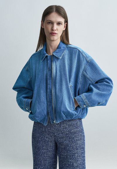 Femme aux cheveux bruns raides portant une veste en jean bleue oversize et un pantalon texturé bleu foncé, mains dans les poches, fond neutre.