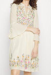 Broderad krämfärgad klänning med klockärmar, med färgglada blommönster och ett midjeband med tofsar. Slätt, lätt material.