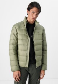 Olijfgroene pufferjack met een gewatteerd ontwerp, hoge kraag en ritssluiting. Draagt over een zwart shirt met een slim fit en een gladde textuur.